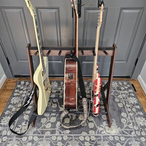 May include: A wooden guitar stand holding three guitars. The guitars include a cream-colored electric guitar, a brown acoustic guitar, and a red and white electric guitar. The stand is on a gray patterned rug.