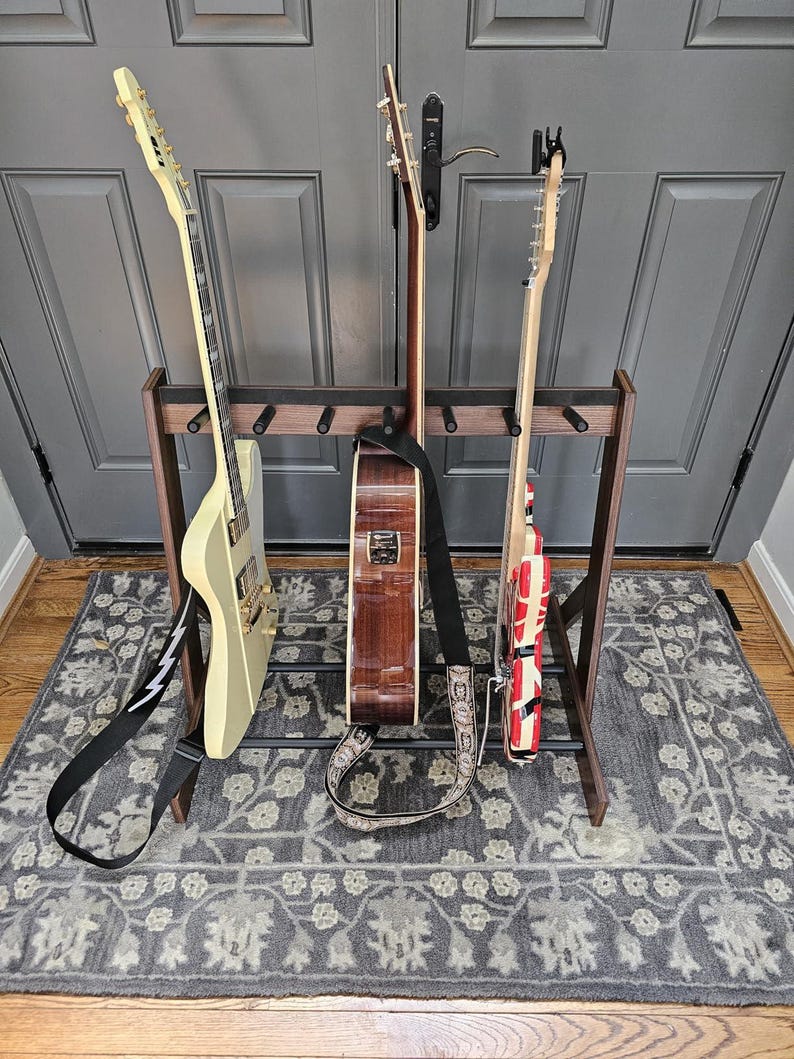 May include: A wooden guitar stand holding three guitars. The guitars include a cream-colored electric guitar, a brown acoustic guitar, and a red and white electric guitar. The stand is on a gray patterned rug.