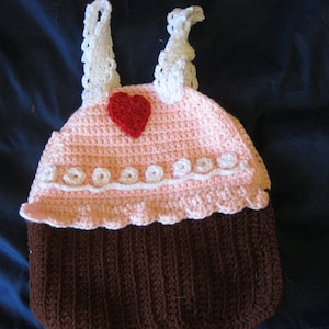 May include: A pink and brown crocheted cupcake hat with white trim and a red heart on top. The hat has two white straps.