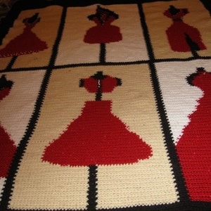May include: A crocheted blanket with a repeating pattern of red dresses on a cream background. The dresses are outlined in black and the background is a cream color.