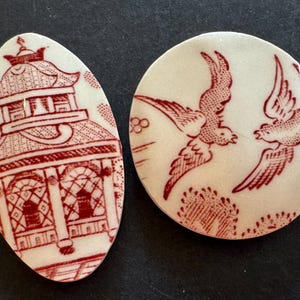 May include: Two ceramic cabochons with a cream-colored base and red transfer-printed designs. One is oval-shaped featuring a pagoda structure, while the other is circular and depicts two birds in flight above stylized foliage and clouds.