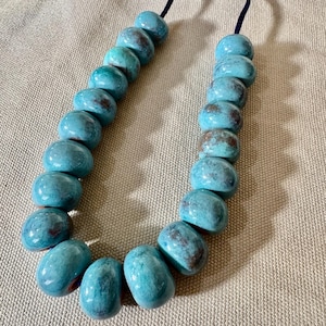 May include: A necklace featuring large, turquoise-colored beads with brown accents. The beads are strung on a dark blue cord, creating a striking contrast. The necklace is displayed on a neutral-toned fabric.