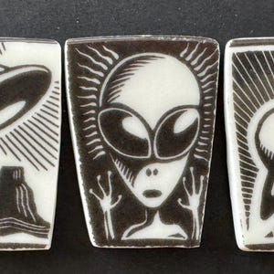 May include: Three rectangular ceramic tiles with black and white illustrations. One tile depicts a UFO over a mountain, another an alien face, and the third a planet with rings. The designs are outlined in black.