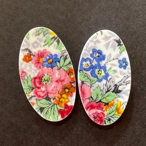 May include: Two oval-shaped ceramic cabochons featuring a vibrant floral pattern. The design includes pink, orange, and blue flowers with green leaves set against a white background with a subtle grey foliage print across the surface.