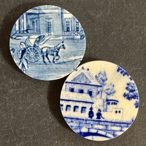 May include: Two circular ceramic discs featuring blue and white transferware patterns. One disc depicts a horse-drawn carriage in front of a building. The other disc shows a building with a bridge and figures, characterized by a visible crackle glaze finish.