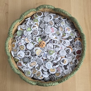 May include: A round, woven basket filled with many small, white buttons. The buttons display cartoon illustrations of ghosts, bats, and pumpkins. The basket has a green trim and rests on a light brown surface.