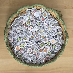 May include: A wicker basket filled with numerous small, circular badges. Each badge displays a colourful illustration of animals, food, and other quirky designs. The basket has a green border and rests on a light wooden surface.