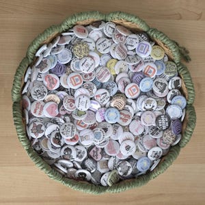 May include: A collection of round badges with various designs, including books and text, fills a woven basket. The basket has a green, braided edge and rests on a light wooden surface. The badges are of various colours.