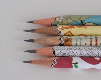 Könnte beinhalten: Fünf Bleistifte mit verschiedenen gemusterten Papierummantelungen. Die Stifte sind vertikal angeordnet, wobei die Spitzen nach rechts zeigen. Die Muster umfassen ein blaues und gelbes Comic-Design, ein gelbes und oranges Kartendesign, ein schwarz-weißes Musterdesign und ein rotes Apfeldesign.
