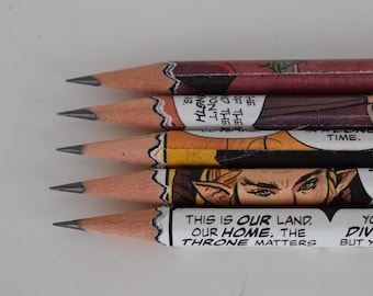 Könnte beinhalten: Fünf Bleistifte mit Comic-Covern. Die Bleistifte sind übereinander gestapelt. Der oberste Bleistift hat den Text "This is our land. Our home. The throne matters." Der unterste Bleistift hat den Text "You have divided but you can..."
