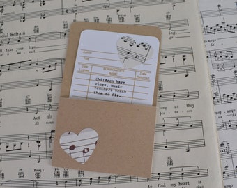 Könnte beinhalten: Ein brauner Papierumschlag mit einer weißen Karte im Inneren. Die Karte hat einen herzförmigen Ausschnitt mit darauf gedruckten Noten. Auf der Karte steht der Text "Kinder haben Flügel, Musik lehrt Lehrer sie fliegen." gedruckt.