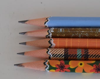 Könnte beinhalten: Nahaufnahme von fünf angespitzten Bleistiften mit dekorativen Designs. Die Bleistifte sind in verschiedenen Farben, darunter Blau, Orange und Grün, mit floralen und gemusterten Designs. Die Bleistifte sind in einer Reihe angeordnet, mit ihren Spitzen nach links.