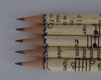 Könnte beinhalten: Fünf Bleistifte mit braunen Holzstiften und schwarzen Graphitspitzen. Die Stifte sind in Papier mit einem Vintage-Notenblattdesign und Text eingewickelt, der "eth comet Wil gome D ver ome dIl evum riv ws t hom osad eslati to I'r" lautet.