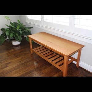 Puede incluir: Un banco de madera con un estante de listones. El banco está hecho de madera marrón claro y tiene un acabado natural. El banco está sentado en un suelo de madera dura.