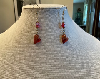 May include: A pair of dangle earrings with a heart-shaped charm and a pink crystal bead. The earrings are made of gold-toned metal.