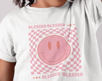 May include: A white t-shirt with a pink smiley face graphic and the text "Blessed Blessed" on a checkered background.