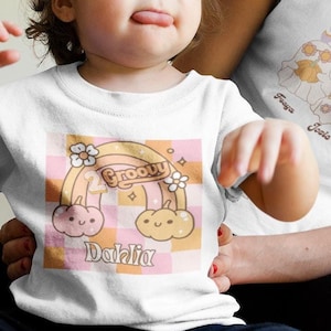 Könnte beinhalten: Ein weißes T-Shirt mit einem Regenbogen und dem Text "Groovy Dahlia" darauf. Ein Kind trägt das Shirt. Eine Frau trägt ein weißes T-Shirt mit dem Text "Boo Crew" und drei Geistern darauf.