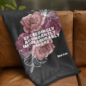 May include: A black throw blanket featuring a floral design in shades of pink and purple. The blanket has the text "EXCEEDINGLY ABUNDANTLY IMMEASURABLY MORE" and "EPH 3:20" in white. The blanket is draped over a brown leather couch.