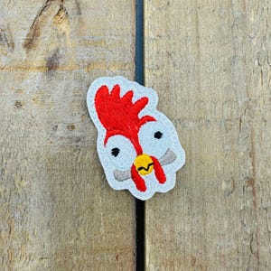 Puede incluir: Parche bordado para planchar con la cara de un gallo con cresta roja, ojos blancos y pico amarillo. El parche está sobre un fondo de madera.