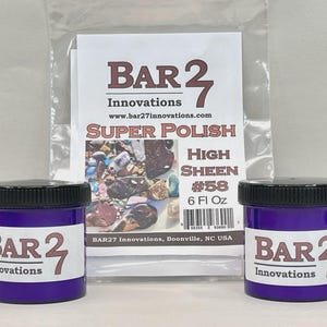 以下が含まれることがあります： 黒いスクリューキャップが付いた2つの紫色のプラスチック製ジャー。それぞれに「Bar 27 Innovations」と書かれた白いラベルが貼られています。ジャーの後ろには、「Super Polish High Sheen #58 6 Fl Oz」と書かれた白い説明書が入った透明なビニール袋があります。