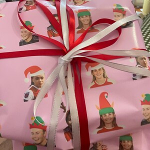 Könnte beinhalten: Eine rosa Geschenkbox, die mit weißem und rotem Band umwickelt ist. Die Box ist mit einem Muster aus Gesichtern verziert, die verschiedene Hüte tragen, darunter Elfenhüte, Weihnachtsmannmützen und Rentiergeweihe.