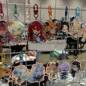 Può includere: Esposizione di ciondoli olografici in acrilico con personaggi in stile anime. Ogni ciondolo ha un design unico con colori vivaci ed è attaccato a un fermaglio in metallo. Il testo "Holographic Acrylic Charms" è visibile.
