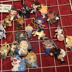 May include: A collection of double-sided character keychains with various designs, each attached to a metal chain. The keychains feature colorful illustrations of anime-style characters. The background is a red surface with a white grid.