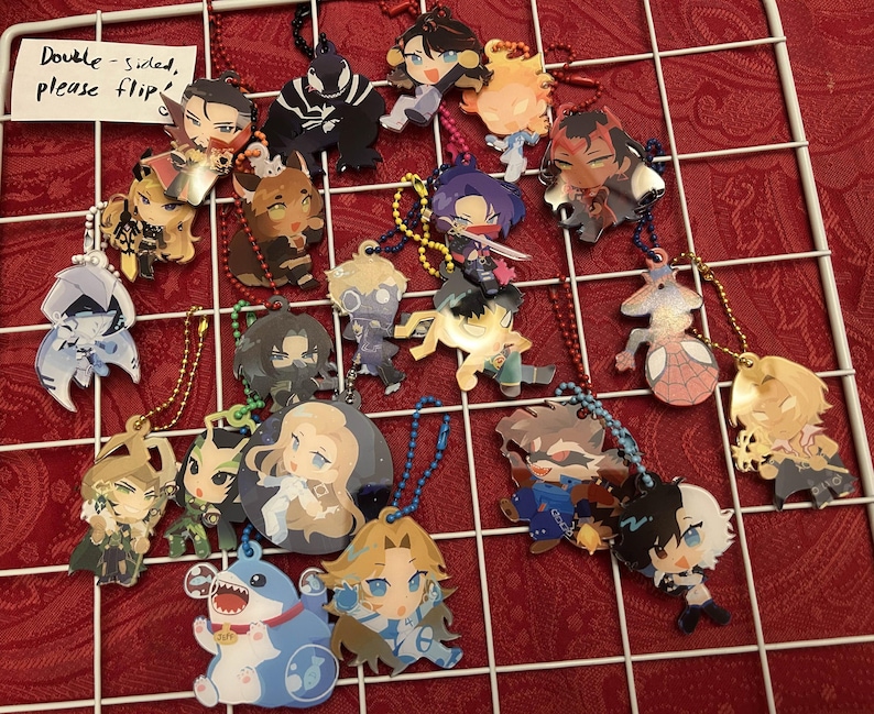 May include: A collection of double-sided character keychains with various designs, each attached to a metal chain. The keychains feature colorful illustrations of anime-style characters. The background is a red surface with a white grid.