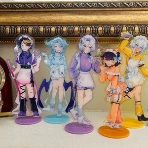 Könnte beinhalten: Eine Sammlung von Anime-Stil-Acrylfiguren mit Charakteren in verschiedenen Outfits, die auf farbigen Sockeln stehen. Die Figuren werden vor einem dekorativen, goldgerahmten Spiegel präsentiert. Ein dekorativer eiförmiger Behälter und eine Uhr sind ebenfalls vorhanden.
