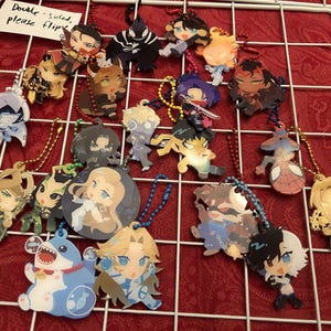 May include: A collection of colorful anime-style character keychains, each with a unique design and attached to a metal chain. The keychains are displayed on a white grid against a red background. The keychains feature various characters.
