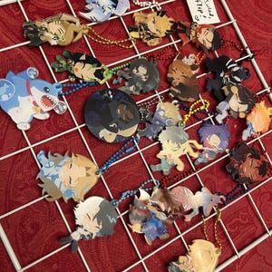 May include: A collection of colorful anime-style character charms with keychains, displayed on a white grid against a red background. The charms feature various designs and are double-sided. Some charms have text that says "Double-sided, Please Flip!"