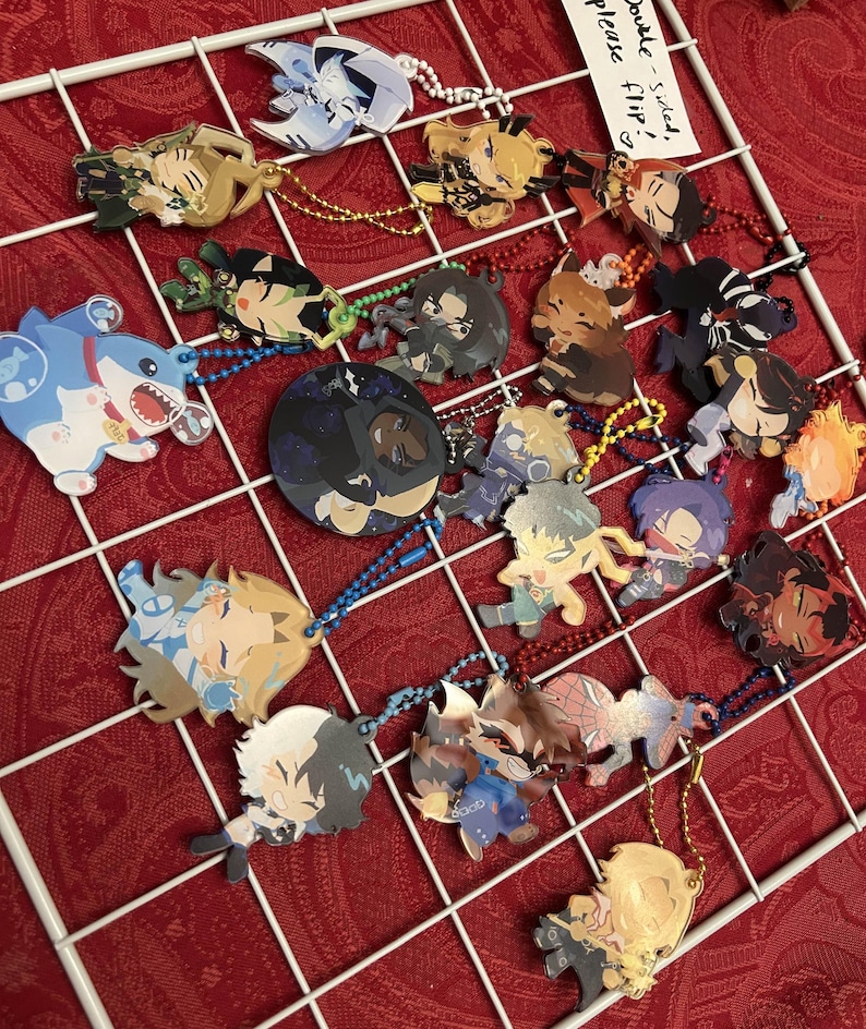 May include: A collection of colorful anime-style character charms with keychains, displayed on a white grid against a red background. The charms feature various designs and are double-sided. Some charms have text that says "Double-sided, Please Flip!"