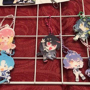 May include: A collection of colorful anime-style character keychains. The keychains feature various characters with different hair colors and outfits. Each keychain is attached to a beaded chain. The keychains are displayed on a white grid against a red background.