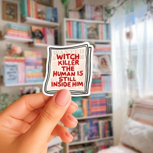 Könnte beinhalten: Ein Aufkleber mit schwarzem Rand und dem Text "WANTED WITCH KILLER THE HUMAN IS STILL INSIDE HIM" in Rot und Schwarz. Der Aufkleber wird vor einem Bücherregal mit Büchern gehalten.