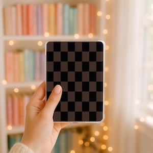 Op de afbeelding: Een doorzichtige telefoonhoes met een bruin en zwart geblokt patroon. De hoes wordt in een hand gehouden, met een vage achtergrond van een boekenplank en lichtslingers.