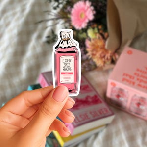 Può includere: Un adesivo rosa a forma di bottiglia di pozione, con l'etichetta "Elixir of Speed Reading". L'adesivo ha un bordo bianco e una parte superiore simile a un tappo. È visibile anche il testo "100%". L'adesivo è tenuto da una persona.