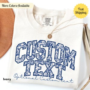 May include: An ivory t-shirt featuring the words CUSTOM TEXT in a large, block-letter font filled with a blue and white floral pattern. Below the block letters, the phrase Optional Custom text is written in a blue cursive script font.
