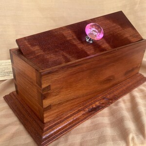 May include: A wooden box with a hinged lid and a pink glass knob. The box is made of dark wood with a natural grain pattern. The box is sitting on a light brown surface.