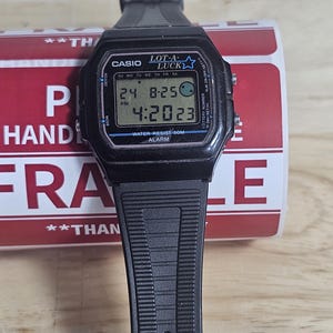 May include: Black Casio digital wristwatch with a black strap. The watch face displays the date, time, and a lottery number. The watch face also has the text "LOT-A-LUCK" and a star.