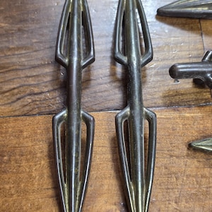 Puede incluir: Primer plano de varias piezas de herrajes decorativos en forma de flecha de color bronce. Las piezas tienen una punta y un diseño estilizado y abierto. Los herrajes son probablemente para muebles o fines decorativos.