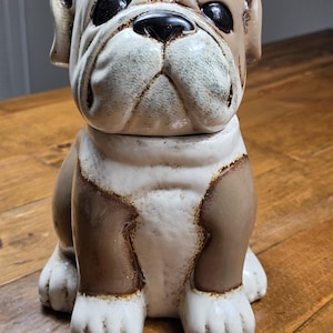 Può includere: Una statuetta di bulldog in ceramica con un mantello bianco e marrone chiaro dalla finitura lucida. Il cane ha il muso rugoso, il naso nero e gli occhi scuri. Il design presenta sfumature marroni intorno alle orecchie, al muso e al petto.