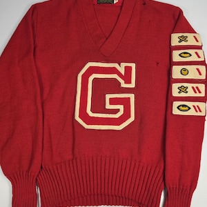 May include: A red V-neck sweater featuring a large cream-colored letter G patch on the chest. The left sleeve displays five rectangular patches, each containing a small yellow football icon and red stripes. The sweater has ribbed cuffs and hem.