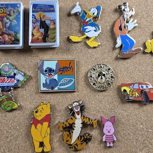 以下が含まれることがあります： ディズニーをテーマにしたエナメルピンのコレクションと、VHS風のミニチュアケース2つ。キャラクターはドナルドダック、グーフィー、プルート、バズ・ライトイヤー、スティッチ、ライトニング・マックィーン、くまのプーさん、ティガー、ピグレット、そして金色のドクロのメダルが含まれています。
