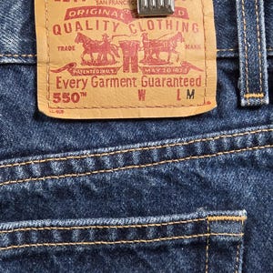 May include: Dark blue denim jeans featuring a tan leather patch on the back waistband. The patch displays the Levi Strauss & Co. logo, two horses, and the text: Levi Strauss & Co. San Francisco Original Riveted Quality Clothing 550 550 W L M.