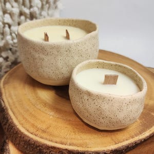Op de afbeelding: Twee gespikkelde keramische kaarshouders met houten lonten. De grotere houder is op de achtergrond en de kleinere houder is op de voorgrond. Beide houders zijn gevuld met witte was en staan op een houten oppervlak.