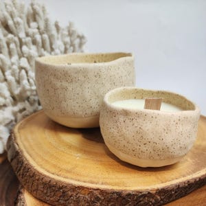Op de afbeelding: Twee gespikkelde keramische kaarshouders met houten lonten. De grotere houder is op de achtergrond en de kleinere houder is op de voorgrond. Beide houders staan op een houten schijf.