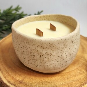Op de afbeelding: Een witte keramische kaars pot met een gespikkelde afwerking. De kaars is lichtgeel van kleur en heeft twee houten lonten. De pot staat op een houten oppervlak.