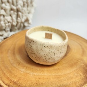 Op de afbeelding: Een gespikkelde witte keramische kaars pot met een houten lont en een witte kaars erin. De pot staat op een houten schijf.