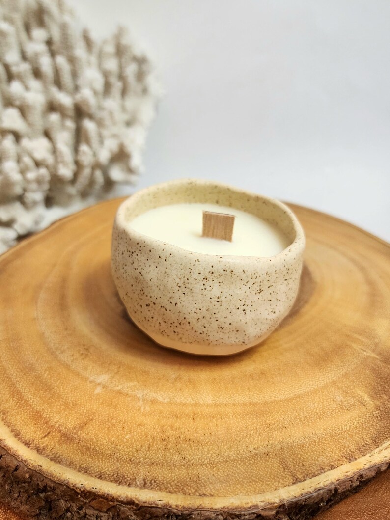 Op de afbeelding: Een gespikkelde witte keramische kaars pot met een houten lont en een witte kaars erin. De pot staat op een houten schijf.