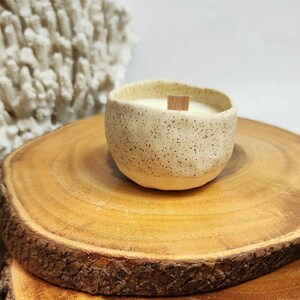 Op de afbeelding: Een gespikkelde witte keramische kaars pot met een houten lont, die op een houten schijf staat.